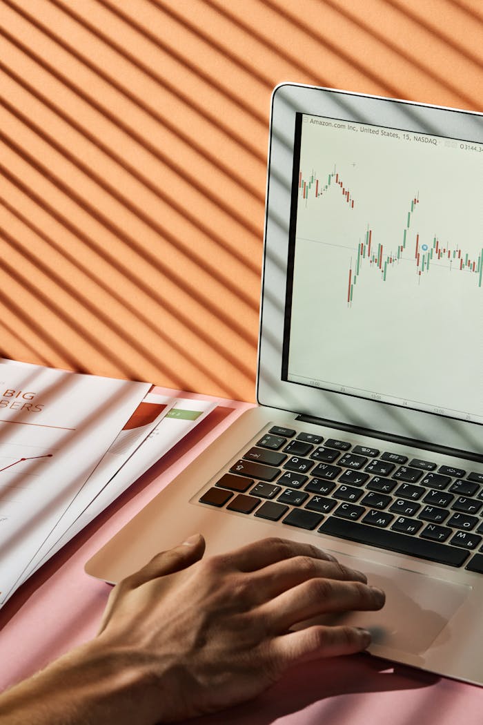 Hand using a laptop with stock market graph, featuring sunlight and shadows across the screen, representing financial analysis.